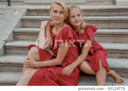 Excited brunette girl embracing with love her mother. Portrait of pretty young woman in dresses. Mother sitting on the steps. Beautiful toddler girl walk with mother, stairs outdoors in the park. 95129778