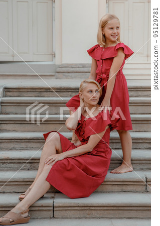 Excited brunette girl embracing with love her mother. Portrait of pretty young woman in dresses. Mother sitting on the steps. Beautiful toddler girl walk with mother, stairs outdoors in the park. 95129781