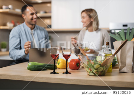 Focus on glasses with red wine. Cheerful multinational couple using laptop eating diet salad Focus on glasses with red wine. Cheerful multinational couple using laptop eating diet salad 95130026