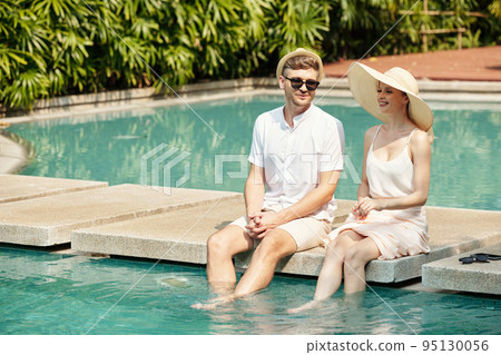 Young couple sitting near the outdoor swimming pool and talking, they spending time at resort Young couple sitting near the outdoor swimming pool and talking, they spending time at resort 95130056