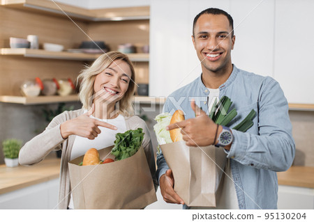 Young arab man and caucasian woman preparing to make salad pointing fingers to each other 95130204