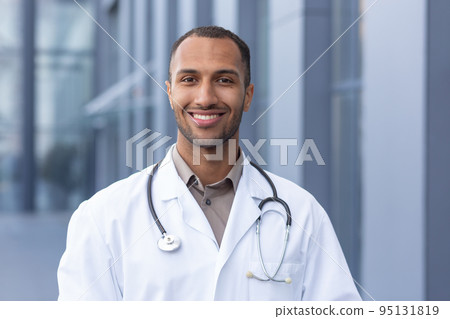 Portrait of successful and smiling African American doctor, man in medical coat with stethoscope looking at camera and smiling, outside modern hospital. 95131819