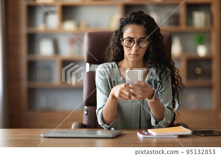 Concentrated woman entrepreneur sitting at workplace, using smartphone Concentrated woman entrepreneur sitting at workplace, using smartphone 95132853