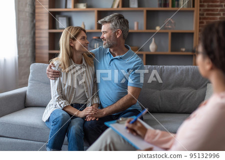 Cheerful black young doctor in glasses looks at middle aged european couple hugging on sofa in clinic Cheerful black young doctor in glasses looks at middle aged european couple hugging on sofa in clinic 95132996