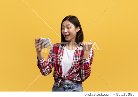 Overjoyed asian lady holding modern smartphone and clenching fist, gambling or trading online over yellow background 95133093