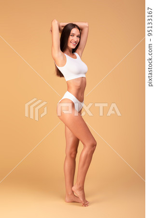 Attractive slender lady with hands up posing in white underwear over beige studio background, full length, vertical shot 95133167