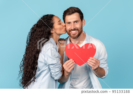 Glad millennial arab female kissing european guy on cheek, holding heart, isolated on blue background Glad millennial arab female kissing european guy on cheek, holding heart, isolated on blue background 95133188