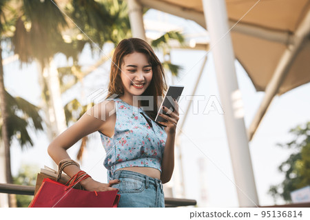 Young adult asian woman using mobile phone for online application and holding shopping bags. 95136814