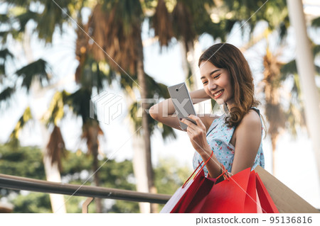 Young adult asian woman using mobile phone for online application and holding shopping bags. 95136816