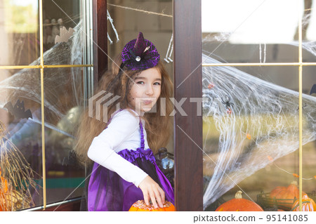 A girl in a witch costume on the windowsill by the window is playing with a spider web and a spider, decorating the house for the Halloween holiday. Terrible scenery, fear and horror, pumpkins 95141085