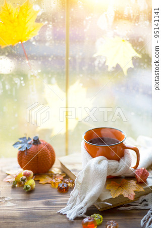 An orange mug in a scarf with hot tea, pumpkins, yellow dry maple leaves, a book on the windowsill, raindrops on the window - autumn mood An orange mug in a scarf with hot tea, pumpkins, yellow dry maple leaves, a book on the windowsill, raindrops on the window - autumn mood 95141141