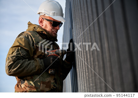 Male builder installing black corrugated iron sheet used as facade of future cottage. Man worker building wooden frame house. Carpentry and construction concept. Male builder installing black corrugated iron sheet used as facade of future cottage. Man worker building wooden frame house. Carpentry and construction concept. 95144117