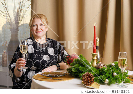 Middle age woman sitting behind the festive table with a glass of sparkling wine in hand Middle age woman sitting behind the festive table with a glass of sparkling wine in hand 95148361
