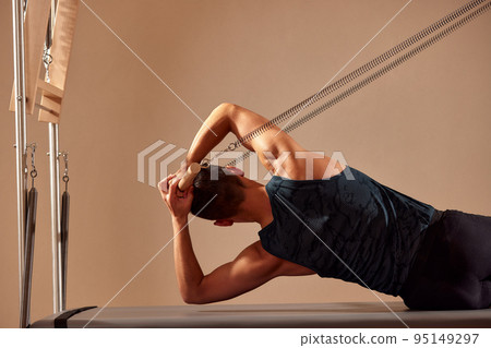 Fit man doing a lunge stretch yoga pilates exercise to strengthen and tone his muscles using a reformer in gym. Health and fitness concept Fit man doing a lunge stretch yoga pilates exercise to strengthen and tone his muscles using a reformer in gym. Health and fitness concept 95149297