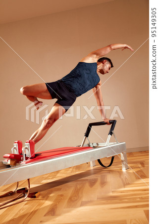 Fit man doing a lunge stretch yoga pilates exercise to strengthen and tone his muscles using a reformer in gym. Health and fitness concept Fit man doing a lunge stretch yoga pilates exercise to strengthen and tone his muscles using a reformer in gym. Health and fitness concept 95149305
