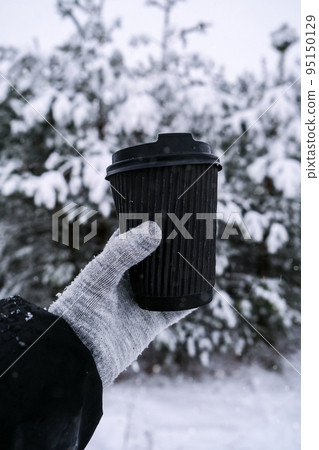 Female hand in winter warm gloves holding black eco paper cup. Winter picnic outdoors. Creative trendy zero waste recycle cup. Mockup. Hot warming drink to go in winter day. Take away concept 95150129