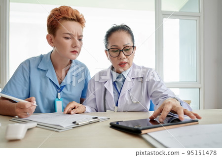 Asian doctor examining image online on digital tablet and showing it to nurse while she filling medical document at table 95151478