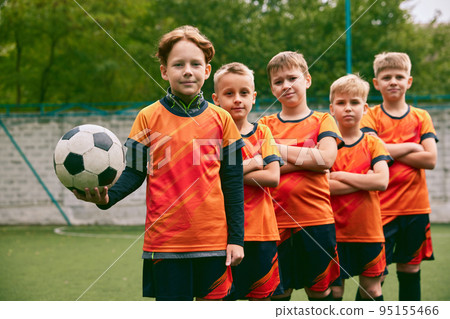 Teammates. Athletic boys in junior soccer team standing together at grass sport field. Football players in orange-black kits and boots. 95155466