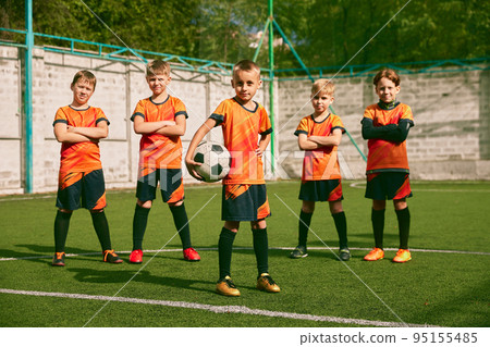Teammates. Athletic boys in junior soccer team standing together at grass sport field. Football players in orange-black kits and boots. 95155485