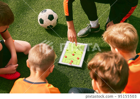 Football training. Soccer coach explaining game rules and strategy using tablet, map. Sports junior team sitting on grass pitch with trainer 95155503