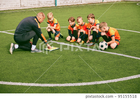 Football training. Soccer coach explaining game rules and strategy using tablet, map. Sports junior team sitting on grass pitch with trainer 95155518