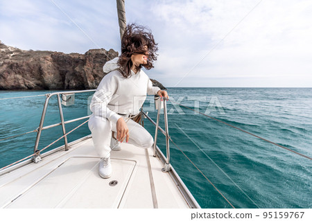 A woman sits on the bow of a yacht on a sunny summer day, the breeze develops her hair, a beautiful sea is in the background A woman sits on the bow of a yacht on a sunny summer day, the breeze develops her hair, a beautiful sea is in the background 95159767
