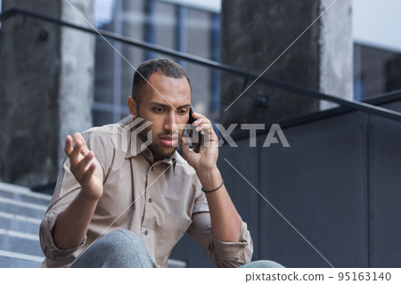 Depressed man sitting on stairs of office building, frustrated talking on phone, upset and fired office worker close up in casual shirt. Depressed man sitting on stairs of office building, frustrated talking on phone, upset and fired office worker close up in casual shirt. 95163140