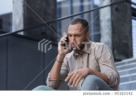 Depressed man sitting on stairs of office building, frustrated talking on phone, upset and fired office worker close up in casual shirt. Depressed man sitting on stairs of office building, frustrated talking on phone, upset and fired office worker close up in casual shirt. 95163142