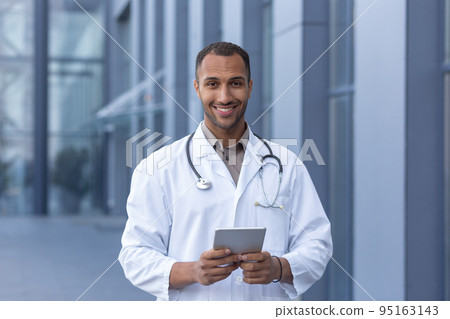 Portrait of happy and smiling hispanic doctor, man smiling and looking at camera, doctor using tablet computer for online consultation, outside of modern clinic. 95163143
