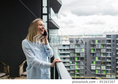 Beautiful blonde woman uses a mobile phone while standing on the balcony of an apartment building Beautiful blonde woman uses a mobile phone while standing on the balcony of an apartment building 95163167