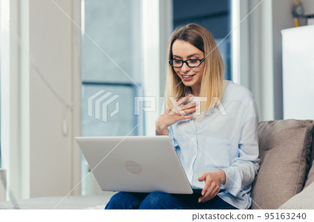 Beautiful blonde woman in casual clothes sitting on a sofa at home, and uses a laptop for video communication with friends and family Beautiful blonde woman in casual clothes sitting on a sofa at home, and uses a laptop for video communication with friends and family 95163240