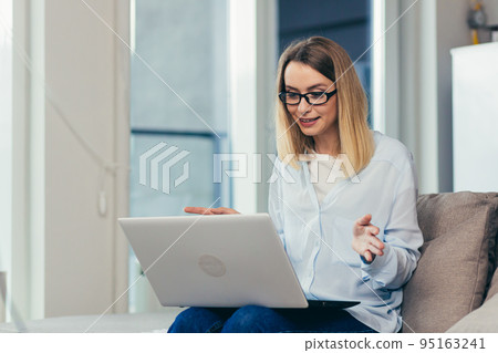 Beautiful blonde woman in casual clothes sitting on a sofa at home, and uses a laptop for video communication with friends and family Beautiful blonde woman in casual clothes sitting on a sofa at home, and uses a laptop for video communication with friends and family 95163241