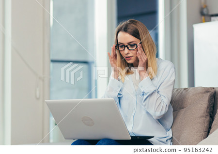 Beautiful blonde woman in casual clothes sitting on a sofa at home, and uses a laptop for video communication with friends and family 95163242