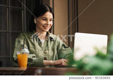 Cheerful Woman Using Earphones and Laptop While Sitting at a Cafe Table, Brunette Girl Working at Restaurant With Notebook and Cocktail. 95163693