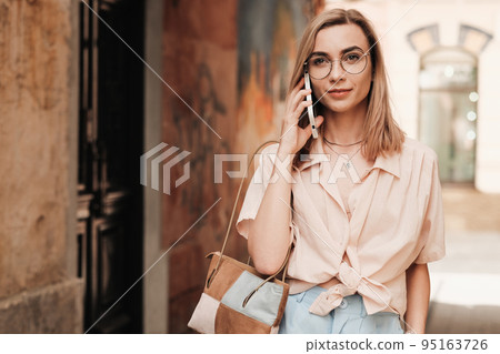 Confident Elegant Woman in Glasses Using Smartphone While Walking in the City, Stylish Blonde Girl with Eyeglasses Standing Outdoors on the Street Having Conversation Through Mobile Phone. Confident Elegant Woman in Glasses Using Smartphone While Walking in the City, Stylish Blonde Girl with Eyeglasses Standing Outdoors on the Street Having Conversation Through Mobile Phone. 95163726