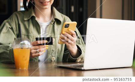 Smiling Unrecognizable Woman Holding Credit Card and Smartphone Making Online Shopping While Sitting at a Cafe Table with Laptop and Cocktail. Smiling Unrecognizable Woman Holding Credit Card and Smartphone Making Online Shopping While Sitting at a Cafe Table with Laptop and Cocktail. 95163755