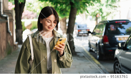 Young Cheerful Woman Using Her Smartphone for Video Call While Walking in the City Street, Brunette Girl Using Cell Phone and Earphones Outdoors. 95163761