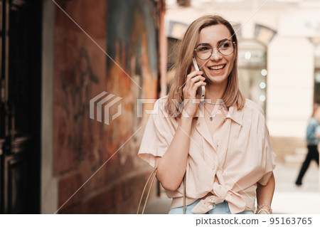 Cheerful Elegant Woman in Eyeglasses Using Smartphone and Smiling, Stylish Blonde Girl with Glasses Standing Outdoors on the Street Having Conversation Through Mobile Phone, Copy Space. 95163765