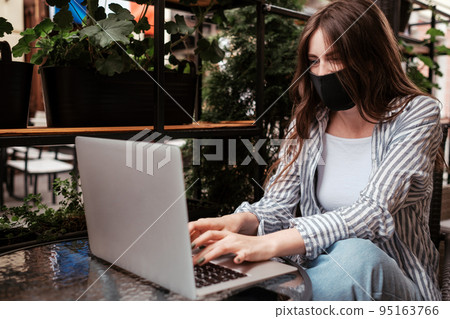 Young Brunette Woman Wearing Protective Face Mask Working on Laptop While Sitting in a Cafe Outdoors, Pandemic Covid-19 Concept Young Brunette Woman Wearing Protective Face Mask Working on Laptop While Sitting in a Cafe Outdoors, Pandemic Covid-19 Concept 95163766
