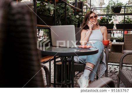 Smiling Young Brunette Woman Talking on Mobile Phone While Sitting with Laptop and Coffee in a Cafe Outdoors Smiling Young Brunette Woman Talking on Mobile Phone While Sitting with Laptop and Coffee in a Cafe Outdoors 95163767