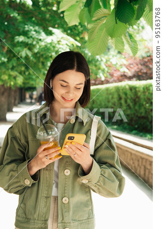 Young Woman Texting on Her Smartphone While Walking in the City Park, Brunette Girl Using Cell Phone and Earphones and Drinking Cocktail. Young Woman Texting on Her Smartphone While Walking in the City Park, Brunette Girl Using Cell Phone and Earphones and Drinking Cocktail. 95163768