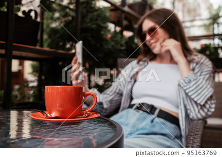 Young Cheerful Woman Using Smartphone While Sitting in a Cafe Outdoors, Focus on Cup of Coffee Young Cheerful Woman Using Smartphone While Sitting in a Cafe Outdoors, Focus on Cup of Coffee 95163769