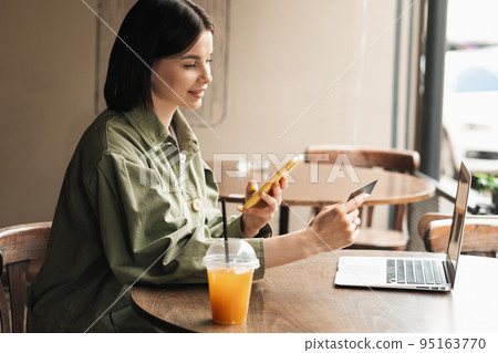 Confident Young Woman Holding Credit Card and Smartphone Making Online Shopping While Sitting at a Cafe Table on Terrace with Laptop and Cocktail. 95163770