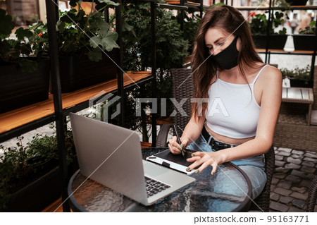 Young Woman Wearing Protective Face Mask Working on Laptop and Drawing Tablet While Sitting in a Cafe Outdoors, Freelance Concept Young Woman Wearing Protective Face Mask Working on Laptop and Drawing Tablet While Sitting in a Cafe Outdoors, Freelance Concept 95163771