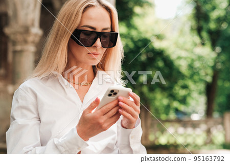 Close Up of Confident Woman Texting on Smartphone While Standing on the Street, Elegant Businesswoman Wearing Sunglasses Looking to the Mobile Phone. Close Up of Confident Woman Texting on Smartphone While Standing on the Street, Elegant Businesswoman Wearing Sunglasses Looking to the Mobile Phone. 95163792