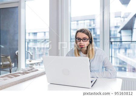 Young blonde woman with a headset and glasses communicates with customers at a distance in front of a laptop or computer screen in the office of an online consumer support call center. conference help 95163812