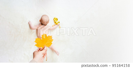 Baby cute child banner. Happy cute baby kid girl lying on white bed background. Mother hand holding yellow leaf. Light background. Little child. Serious emotion. 95164158