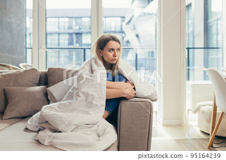 Young woman covered with a blanket sitting on the couch at home, in a bad mood, tired, offended, looking sadly at the camera 95164239