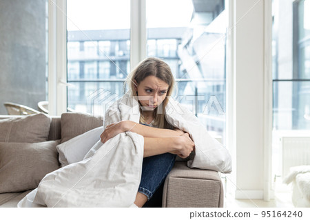 Young woman covered with a blanket sitting on the couch at home, in a bad mood, tired, offended, looking sadly at the camera 95164240