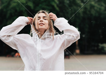 Close Up Portrait of Young Woman Wearing Raincoat While Standing on the Street at Rainy Day, Blonde Girl Dressed in Waterproof Cloak Having Fun Under Rain in City. 95164704